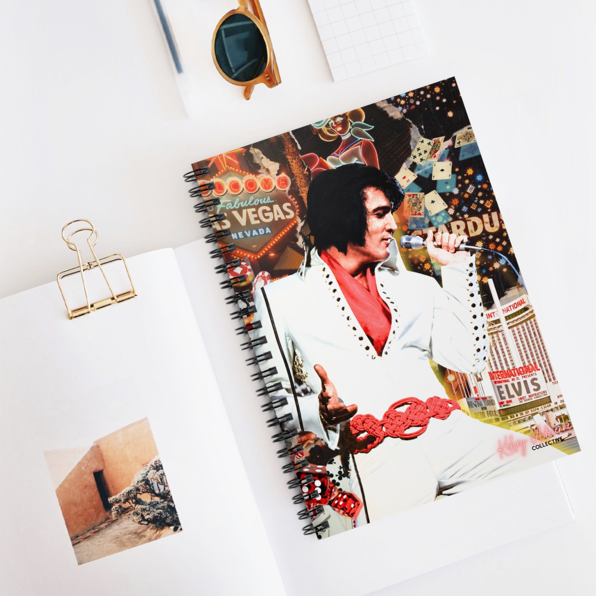 A spiral-bound notebook featuring a collage print of Elvis Presley in a Las Vegas themed design, styled on a white desk with an open book, a gold binder clip, and sunglasses.